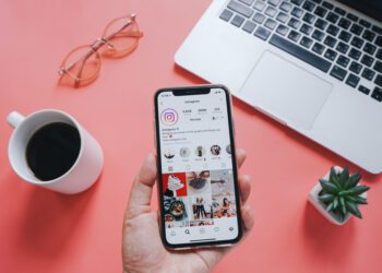 Hand holding Apple iPhone XS with Instagram application on the screen with laptop and coffee background. Instagram is a photo-sharing app for smartphones.